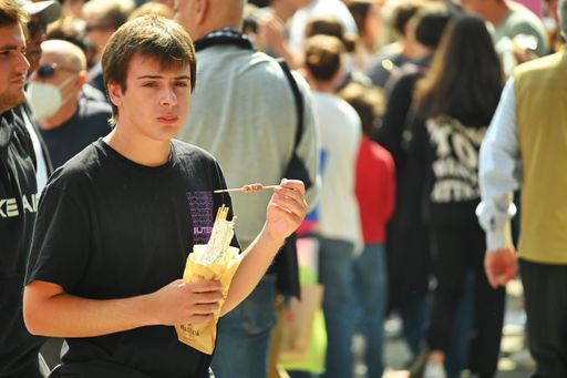 A boy walking in a crowded area while eating Italian street food on a sunny day