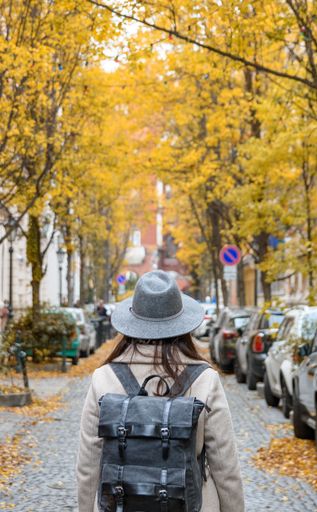 A vertical shot of a woman in a gray hat and coat walking in the urban street with yellow autumn trees