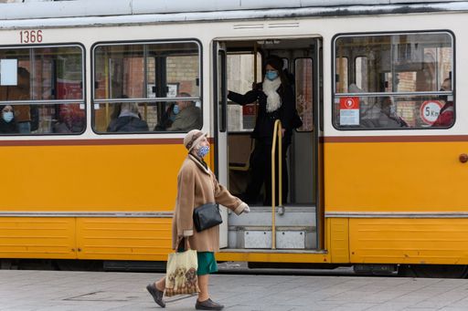 An elderly woman wearing a face mask and walking on the sidewalk in front of the yellow tram