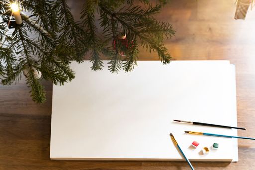 A stack of blank white paper with paint and brushes on it under a Christmas tree - copy space for text or drawings