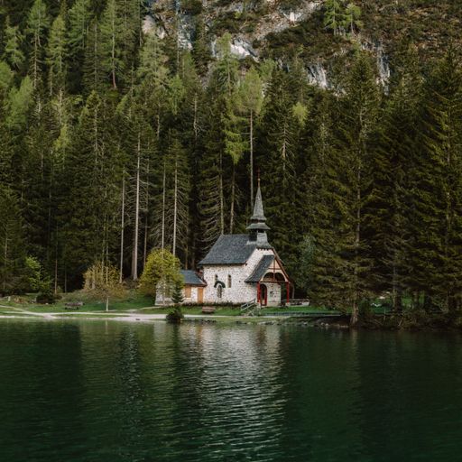 A beautiful shot of an old chapel against green pine trees reflecting on Lake Braies in Italy