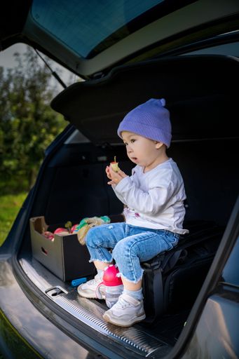 An Asian toddler sitting on a bag in a car trunk with picked apples enjoying the fruit