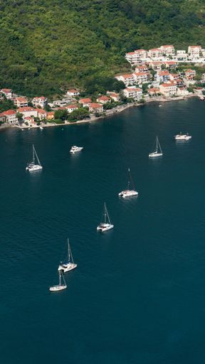 A vertical aerial shot of white boats sailing in the ocean along the coast in daylight