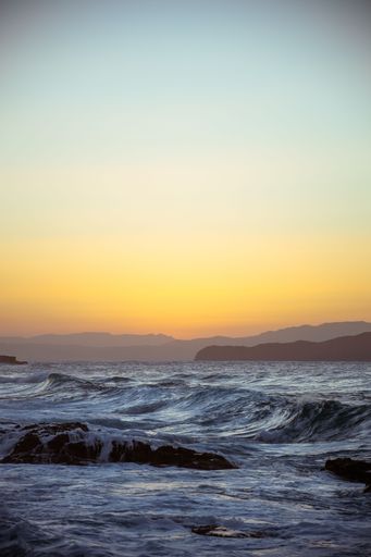A vertical shot of sea waves on the shore with mountain silhouettes on the horizon during sunset