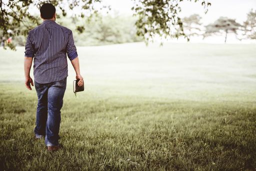 A male walking on a grassy field while holding the bible with a blurred background shot from behind
