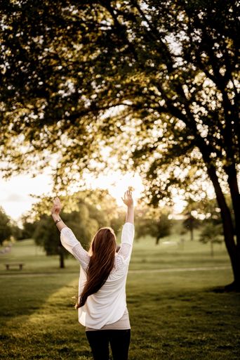 A vertical shot from behind of a female standing in a grassy field with her hands up and a blurred background