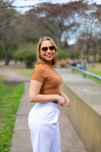A Young Hispanic female in brown top and white pants smiling on camera in the park