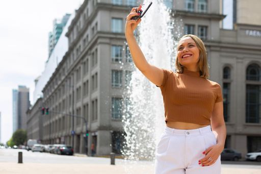 A Young Hispanic female in brown top and white pants taking selfie ina street