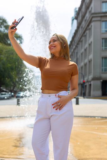A Young Hispanic female in brown top and white pants taking selfie in a street
