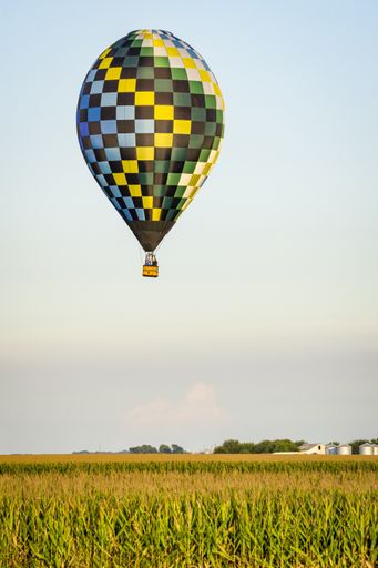 Vertical image of checkered hot air balloon with passengers in gondola flies over cornfield clouds, white farm in distance.