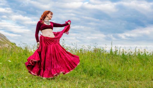 Young beautiful and happy red-haired girl dances incendiary dance in red ball gown on areen meadow against blue cloudy summer sky.