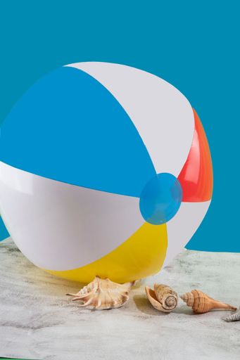A vertical shot of a beach ball with red, yellow, blue, and white stripes with various shells next to it on a blue background
