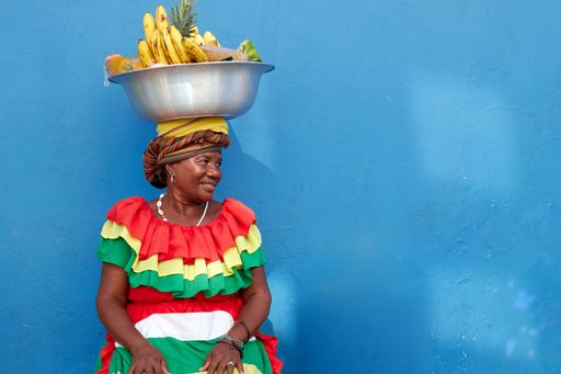 A regular footage of a Palenquera holding a big bowl of fruits above head, dressed with colorful clothes on a blue background in Cartagena, Spain