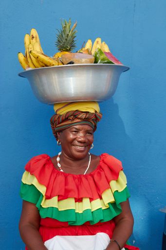 A vertical footage of a Palenquera holding a big bowl of fruits above head, dressed with colorful clothes on a blue background in Cartagena, Spain