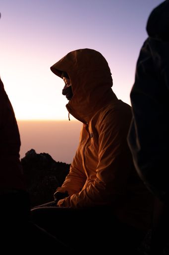 A young adult male stands atop a mountain, dressed in a hooded sweatshirt
