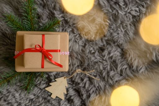 A top view of a Christmas gift box with a red ribbon, blur lights, and pine branches on a cozy gray carpet