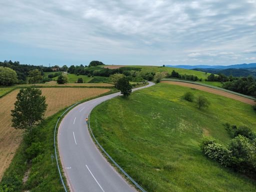 An aerial view of a narrow road surrounded by green fields and mountains against a blue cloudy sky