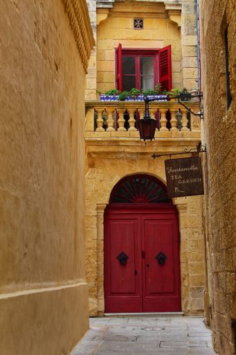 A vertical shot of an alleyway leading to an old building with a red door and a cute balcony in Mdina, Malta