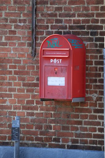 A vibrant red postbox mounted against a brick wall adjacent to a parking meter, illuminated by natural light