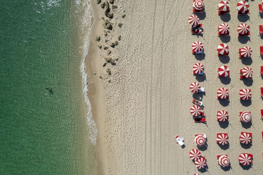 A symmetric aerial view of the beach with people resting under the red and white umbrellas on the shore