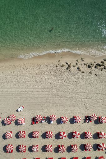 A symmetric aerial view of the beach with people resting under the red and white umbrellas on the shore