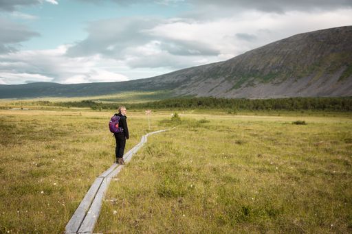 A Caucasian woman with backpack walking on a trail in a green field with mountain in the background