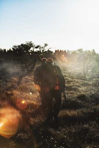 A vertical shot of two male hunters with guns walking in the forest under the sunlight