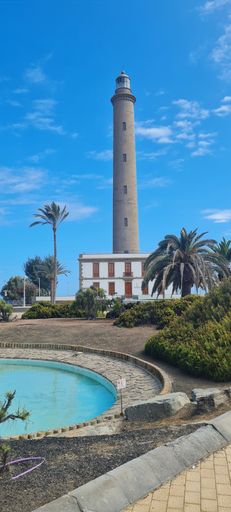 A vertical shot of a tall lighthouse surrounded by palm trees under a bright blue sky in Maspalomas, Spain