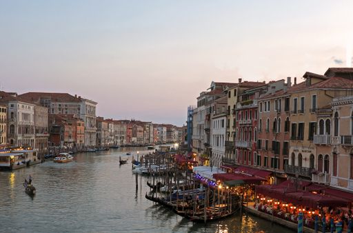 The Grand Canal in Venice, Italy, at dusk, bathed in the golden glow of the setting sun, viewed from the Rialto Bridge. Also in image: people, boats.