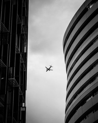 A vertical shot of the silhouette of an airplane visible between two modern skyscrapers under a gloomy gray sky