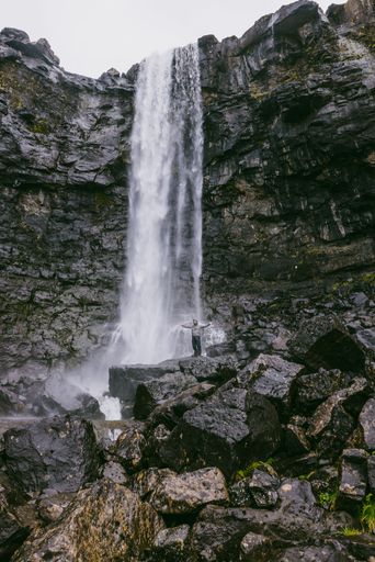A vertical shot of a man standing on rocks with the background of the Fossa waterfall in the Faroe Islands