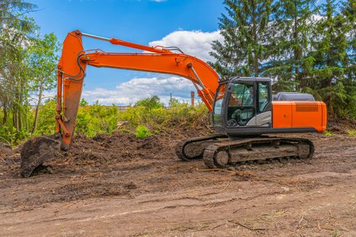 Large orange crawler excavator stands against backdrop of agricultural field with trees and blue cloudy summer sky.