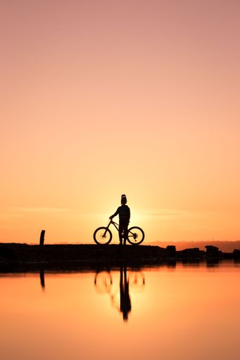 A vertical of cyclist silhouette near a pond at sunset with water reflection