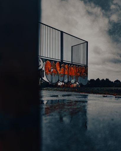 A vertical shot of colorful graffiti on a wall below a fence in a skatepark with puddles on the ground