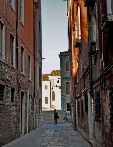 Typical narrow pedestrian street in Venice, Italy, showing exteriors of old houses and unrecognisable female walking away from camera towards bridge.