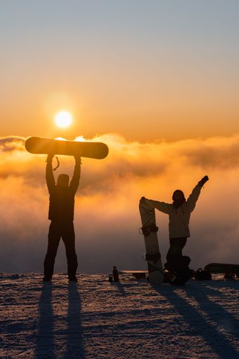 Two snowboarders in silhouette enjoying the winter landscape from the top of a mountain, holding their boards in anticipation of next exhilarating run