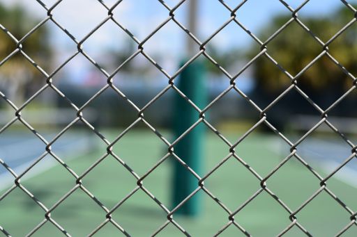 A closeup of a metal mesh with blurry tennis courts and a green pole in the background.