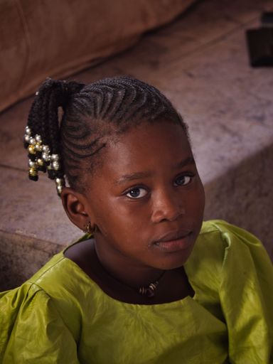 A portrait of an African kid with beautiful hairstyle sitting on the floor inside a house, vertical