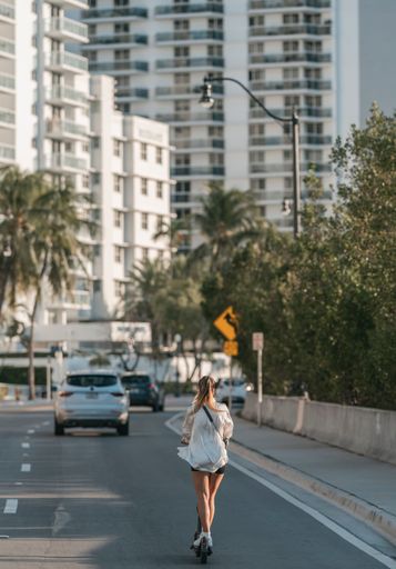 A Vertical shot of a girl on a scooter in front of white buildings and trees on the side of the road in the daytime