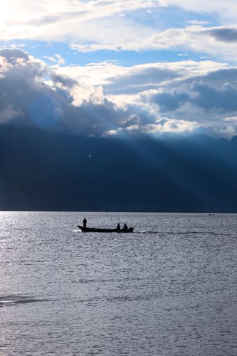 A vertical shot of the silhouette of a boat with people against the background of blue cloudy sky.