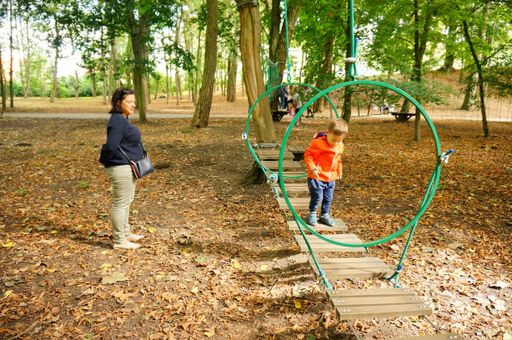 Small three years old child walking on a wooden bridge of a obstacle route while mother is watching at a park with trees.