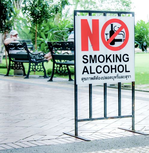 A park with a sign in Bangkok, with No smoking alcohol written in red and black letters on a white background. A person is sitting down on a bench.