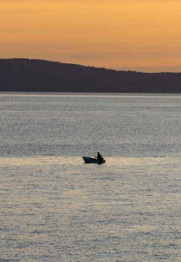 A silhouette of a fisherman on the boat in the middle of a lake with a hill forest in the background under a sunset sky