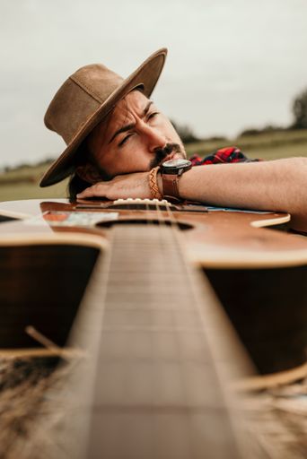 A vertical shot of a young man with a brown hat posing with a guitar in a rural setting