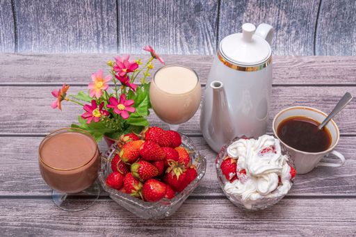 On wooden table there are vases with ripe strawberries, teapot, glasses and wine glasses with drinks. Still life with positive emotions.