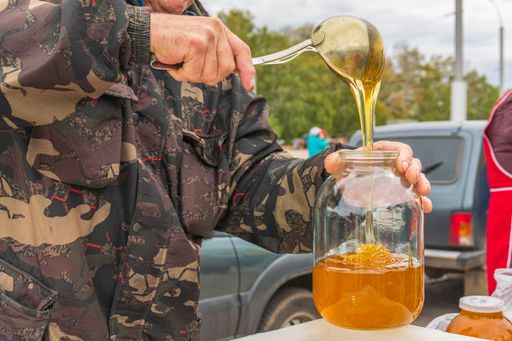 Autumn agricultural fair. Behind counter, man pours bee honey into glass jar using ladle. Preparing goods for sale.