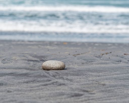 A single large stone alone on a sandy beach with an ocean wave in the background.