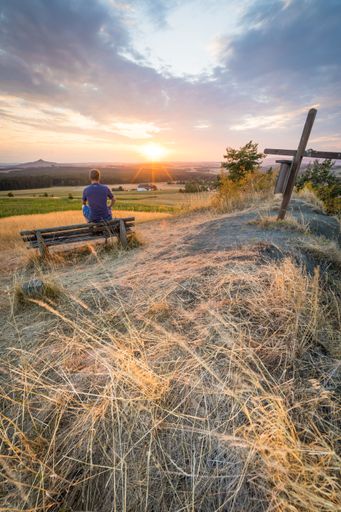 A man sitting on a bench in a wheat field enjoying the view of the beautiful sunset