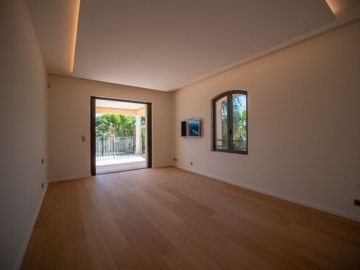 A large modern room with wooden floor, light beige walls, and a window opening onto a balcony