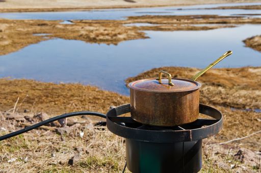 A large dirty round pot with a lid being cooked on black outdoor stove on the lakeshore